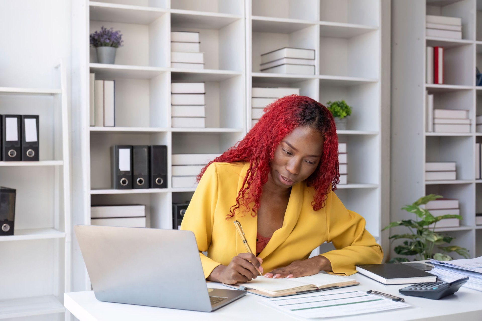 Jeune femme d’affaires afro-américaine en blazer jaune assise à un bureau faisant de la paperasse dans un cadre de bureau moderne